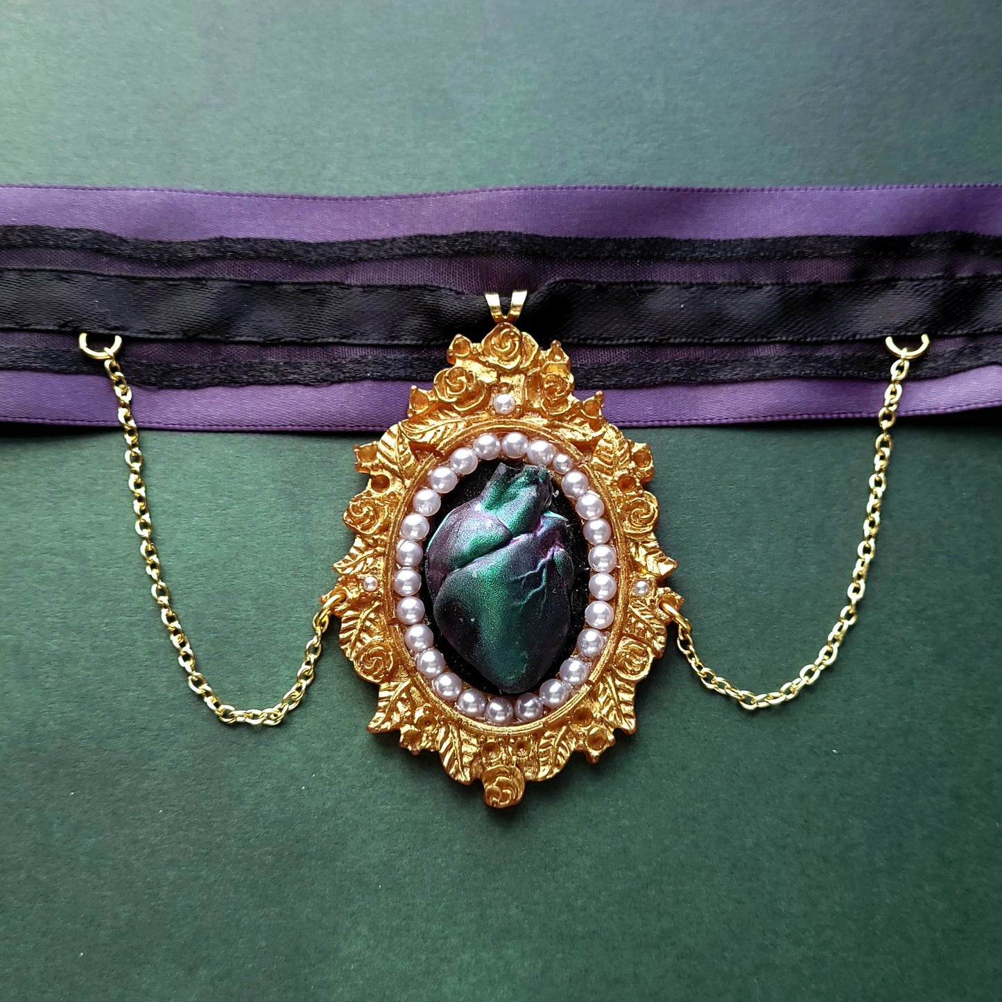 A gold frame pendant with pearl details and green purple shifting heart in the center. Two gold chains dangle off the sides and connect to a black ribbon layered on a striped ribbon then on purple.