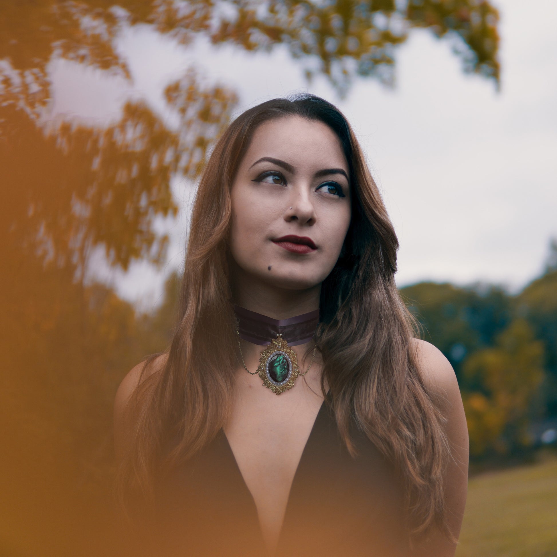 A model with long brown hair looks off into the distance while wearing a purple choker with a hanging gold pendant. The pendant has pearls and a green heart inside.